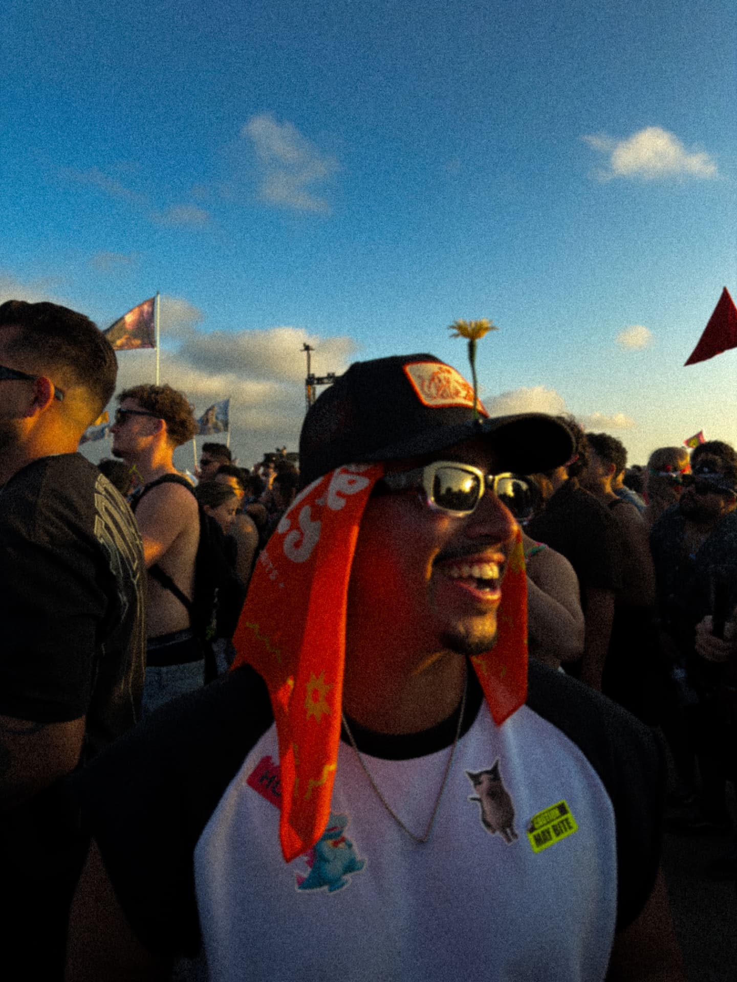 Smiling face in the festival crowd at golden hour — cap with a flower tucked in, orange bandana, OBSDYN frames catching the last light